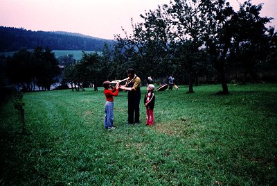 Vintage family moment in a lush orchard, likely mid-20th century. Adult in striped shirt teaches two children (one in red, on...