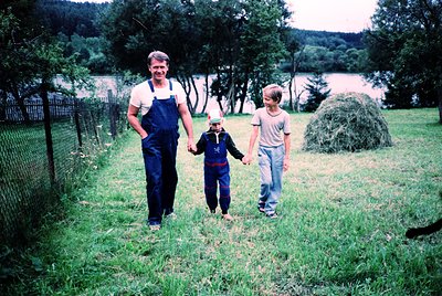 A family of three walks hand-in-hand across a rural meadow, framed by a chain-link fence and haystacks. The man wears overall...