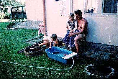 Vintage backyard scene with family playing in a blue plastic kiddie pool filled via garden hose. Adults and children dressed ...