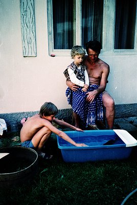 Family plays in a blue plastic tub on a grassy courtyard, likely mid-20th century. Adult wears patterned shorts, child in pol...