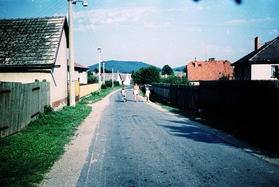 Vintage street scene with three children playing on a narrow, paved road flanked by modest homes and fences. Distant mountain...
