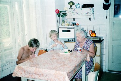 Three generations share a meal in a mid-century kitchen, 1970s Eastern Europe. White-tiled walls, floral-patterned tablecloth...