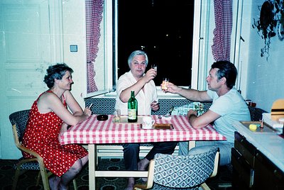 Three adults toasting indoors, 1970s European home. Woman in red polka-dot dress, man in white shirt raising glass, another m...