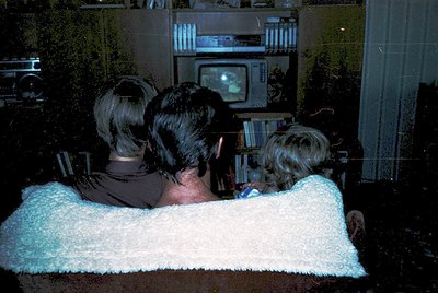 Vintage CRT TV in dimly lit living room with three seated figures watching. Mid-century furniture, vinyl record player, and b...
