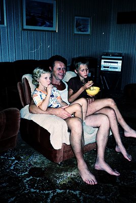 Family trio in a mid-20th-century living room, likely 1960s–1970s. Woman in floral dress and man in casual shirt share a sofa...