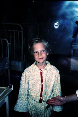 Vintage black-and-white photo of a young boy in a floral-patterned robe, standing beside a hospital bed with metal railings. ...