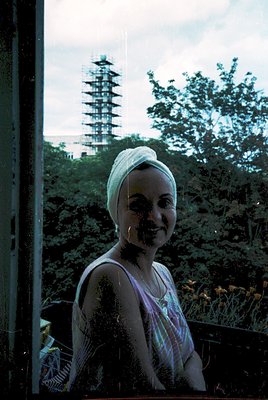 Vintage photo of a woman in a wet, patterned dress and headscarf, framed by a window. Behind her, a pagoda-like tower rises t...