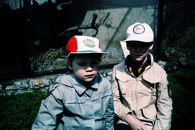 Two boys in outdoor zoo attire, 1970s-1980s. Left wears a red-and-white "Castrol" cap with matching jacket; right sports a wh...