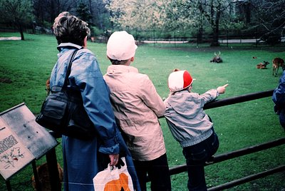 Three figures in a park setting, mid-20th century. Adults in coats and hats, child in a red-and-white striped cap, pointing t...