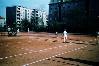 Vintage clay-court tennis match in urban setting, likely 1970s. Players in retro attire (shorts, knee-length skirts) on a mar...