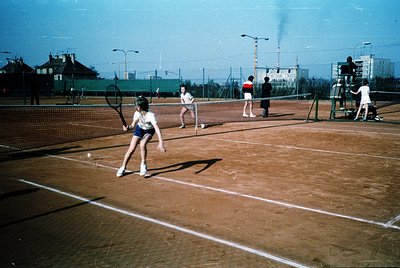 Vintage tennis court with clay surface, featuring a young player mid-forehand shot. Six individuals—four active, two observin...