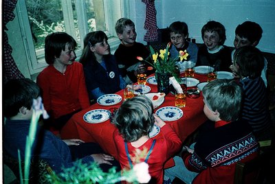 Family gathering at a round table in a cozy indoor setting, likely a 1970s-1980s home. Blue-and-white floral tableware, yello...