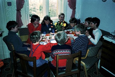 Family gathering in a cozy 1970s dining room, featuring floral-patterned curtains and vintage chairs. Centerpiece includes ye...