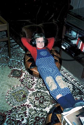 Vintage 1970s interior shot of a child reclining on a patterned rug with headphones, wearing a polka-dot sweater and knee-hig...