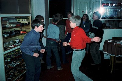 Vintage indoor gathering of young boys playing a game with toy cars on a table, lit by a single overhead bulb. Shelves displa...