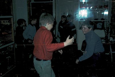 Vintage arcade scene with two players engaged in a competitive game, likely *Pong* or early pinball. Reflections in glass cre...