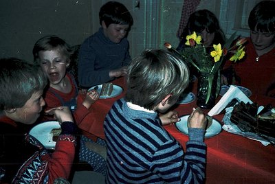 Vintage indoor gathering of children in 1970s/80s attire, seated at a round table with plates, cups, and a vase of yellow flo...