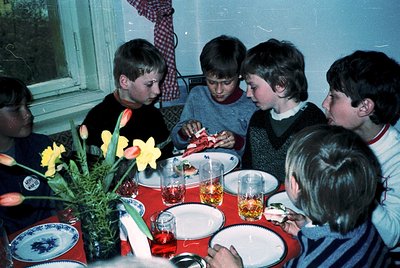 Five boys seated around a round table in a cozy indoor setting, likely a 1970s-1980s family gathering. Table adorned with blu...