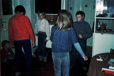Vintage indoor scene featuring four children in 1970s-era clothing—sweaters, jeans, and button-ups—standing in a dimly lit ki...