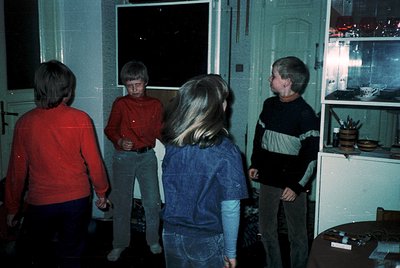 Four children in 1970s-era clothing gather indoors, likely a home or community space. The boy in red stands near a window, wh...