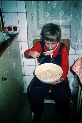 Child in 1970s-era kitchen holding a bowl of batter, spooning mixture into a pan. White-tiled walls with floral-patterned wal...