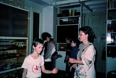 Vintage indoor scene featuring a family in a retro kitchen, likely 1970s–1980s. A woman in a floral blouse stands beside a bo...