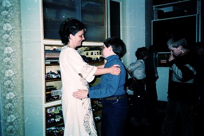 A woman in a patterned blouse hugs a young boy in a blue sweater inside a retro arcade, surrounded by classic pinball machine...