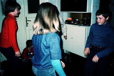 Vintage indoor scene of four children playing in a dimly lit kitchen, likely late 1970s–early 1980s. White cabinetry, vinyl f...