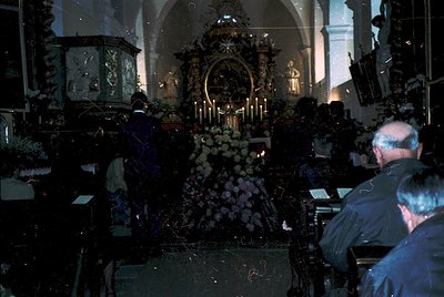 Ornate Orthodox church interior with candlelit tombs draped in floral tributes. Iconostasis features intricate gold and stain...