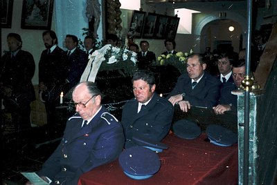 Formal funeral ceremony featuring three men in military uniforms (blue with insignia) seated at a draped table with floral ar...