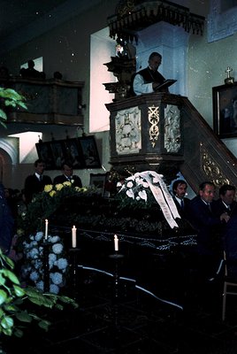 Religious funeral service in a dimly lit church, featuring a decorated casket draped in white flowers and candles. A priest s...