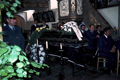 A solemn funeral procession inside a church, featuring a decorated casket draped in flowers and candles. Attendees in formal ...