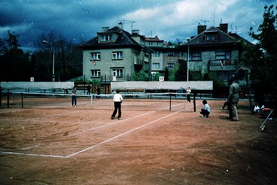 Vintage outdoor tennis court with clay surface, marked for doubles play. Residential buildings with gabled roofs and satellit...