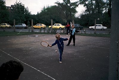 A young child in a blue tracksuit mid-tennis swing on a concrete court, 1970s urban park. Vintage cars (yellow, white, red) p...