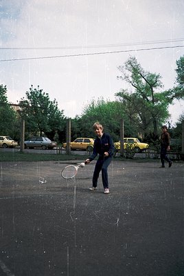 Vintage tennis scene in urban park, 1960s-70s. Woman mid-serve on asphalt court, wearing dark jacket, jeans, and sneakers. Ye...
