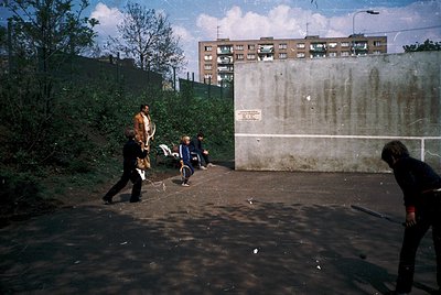 Vintage urban street scene with three individuals playing frisbee in a paved area. Concrete wall with faded signage ("ПОЧТА С...