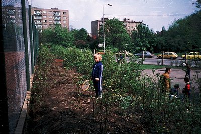 Vintage urban scene featuring a child in a striped sweater holding a tennis racket amidst overgrown vegetation. Mid-20th-cent...