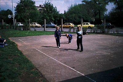 Vintage outdoor bocce court with chalked lanes, surrounded by parked vintage cars (, , ). Uniformed officials and players in ...