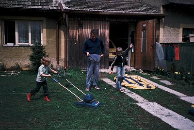 Mid-20th century suburban scene: adult supervises two children operating lawn mowers in a residential yard. Yellow inflatable...