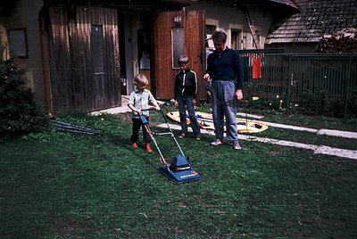 Three children and an adult mow lawn in suburban backyard, 1970s. Wooden garage door, wooden fence, and vintage push mower do...