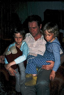 Family portrait featuring a man and two children seated indoors, likely mid-20th century. The man wears a light-colored, patt...