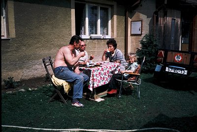 Three adults seated outdoors on simple wooden chairs around a small table, sharing a meal in a residential courtyard. The man...
