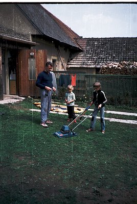 Family mows lawn in rural backyard, 1970s. Man in dark sweater and jeans directs two children operating vintage reel mowers. ...