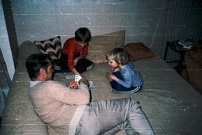 Three individuals play cards on tiled floor in dimly lit room with concrete walls. Adult in beige sweater and trousers sits c...