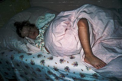 Vintage black-and-white photo of a pregnant individual resting on a floral-patterned bedspread, likely mid-20th century. Soft...