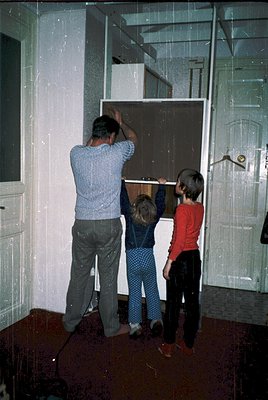 Mid-20th century family installing a built-in fridge in a retro kitchen. Adult male in striped shirt and jeans assists two ch...