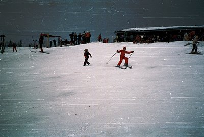 Vintage ski slope with groomed snow and wooden structures, likely a 1960s-70s alpine resort. Two skiers—one adult in bright o...