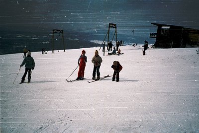 Vintage ski slope scene with four skiers in mid-20th-century gear, posing on groomed snow. Wooden ski lift poles and a small ...