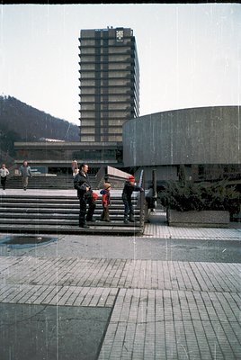 Modernist brutalist architecture with a stepped, concrete high-rise and angular plaza. Three figures—two adults and a child—s...