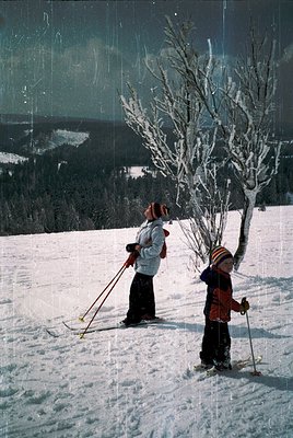 Two skiers pose on a snow-covered slope, likely a mid-20th century ski resort. The adult wears classic 1960s ski gear (white ...